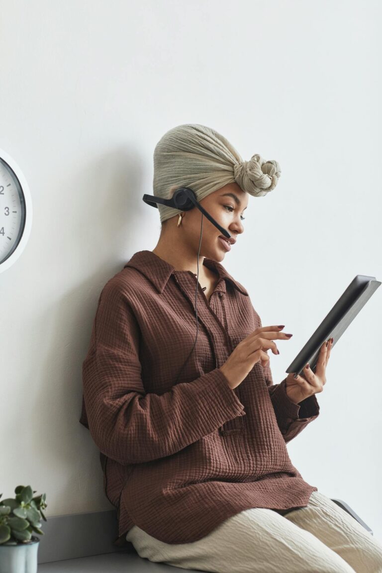 Focused African American female call agent with microphone browsing tablet while sitting at white wall during work in light room