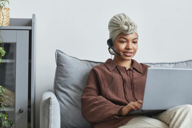 Focused African American female call agent with handsfree headset typing on netbook while sitting on couch near cupboard in light room